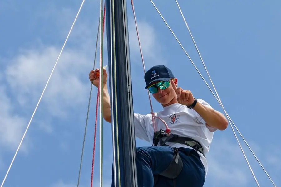 A sailor part way up the mast, harnessed on, working the rigging